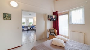 Een slaapkamer met een bed, een televisie, een rieten stoel en uitzicht op een zithoek.