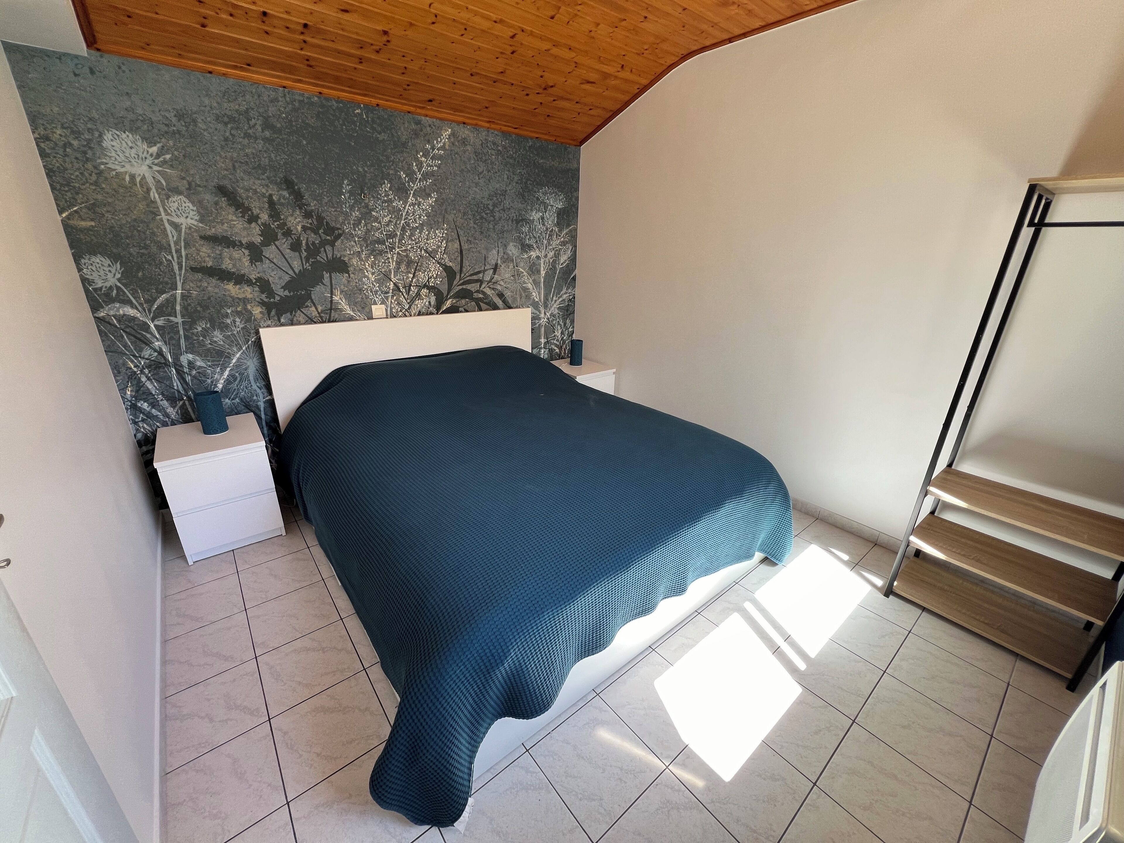 A bedroom with a bed, white nightstands, a wooden headboard, and a wall with a floral pattern.