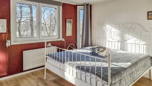 Une chambre avec un lit en métal, un radiateur, une fenêtre donnant sur des arbres sans feuilles et un mur blanc orné d’un cadre.