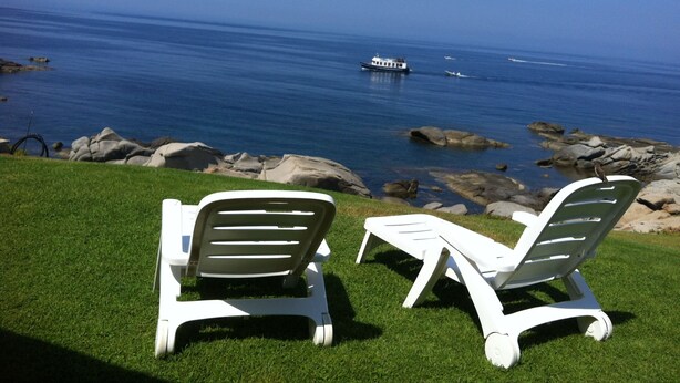 Two white lounge chairs on a grassy area overlooking a rocky shoreline with a boat in the water. - Panoramic villa on the sea Villasimius (Villasimius)
