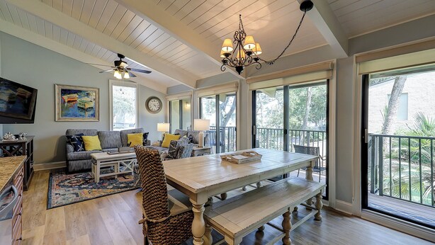 A spacious living room with a large wooden table, comfortable seating, a ceiling fan, and large windows offering a view of the outdoors.