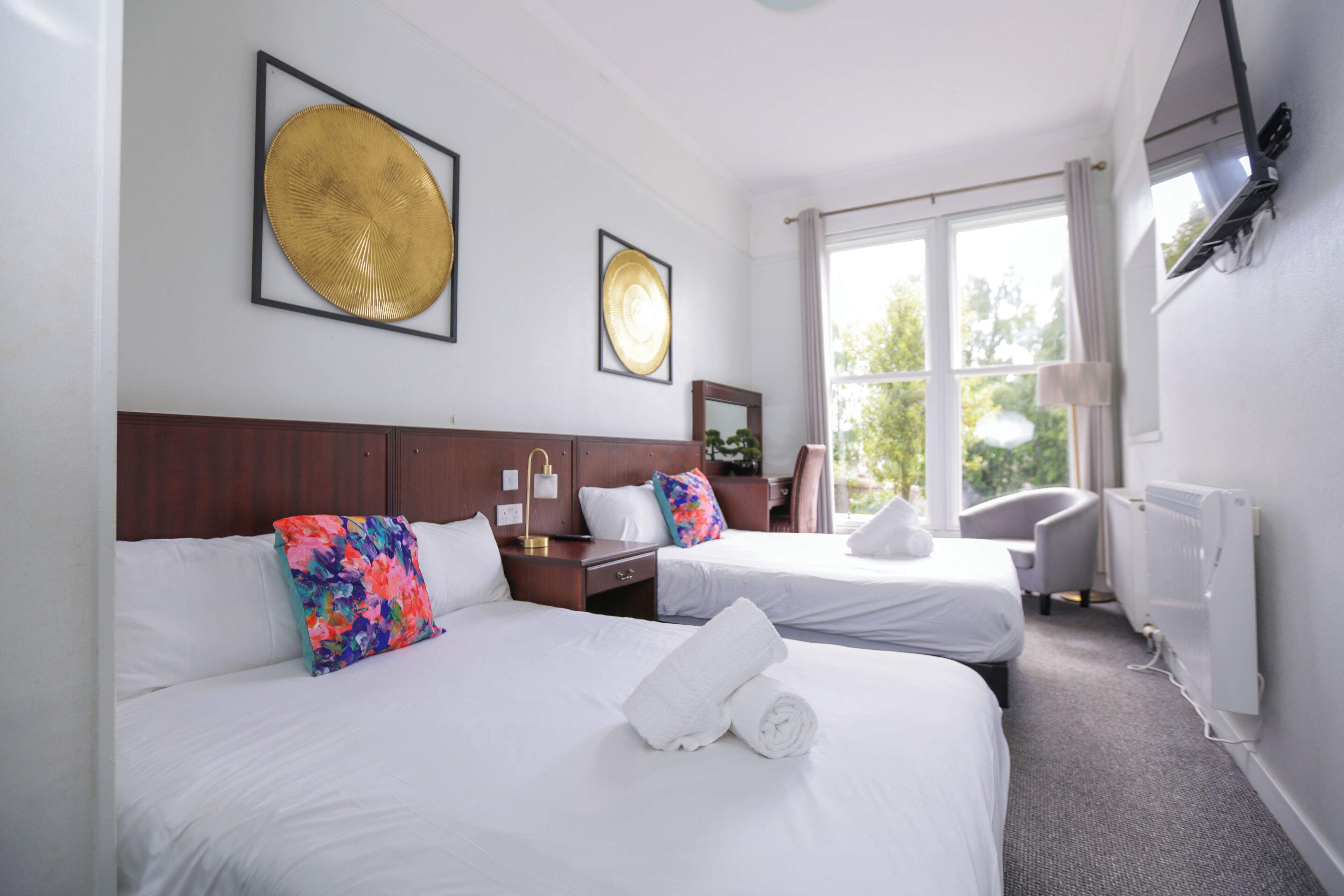 A hotel room with two beds, a wooden headboard, a desk with a chair, a radiator, and a window with curtains.