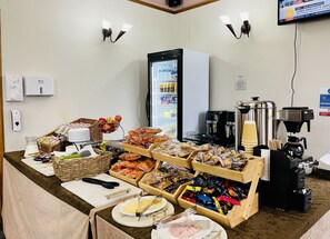 Montaje de bufé con diversos alimentos, un refrigerador, máquinas de café y un televisor mostrando noticias.