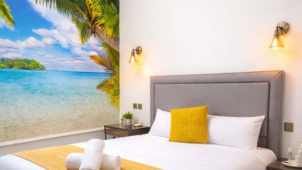 Una cama bien hecha con un cabecero gris, ropa de cama blanca y amarilla, y un mural de playa tropical en la pared.
