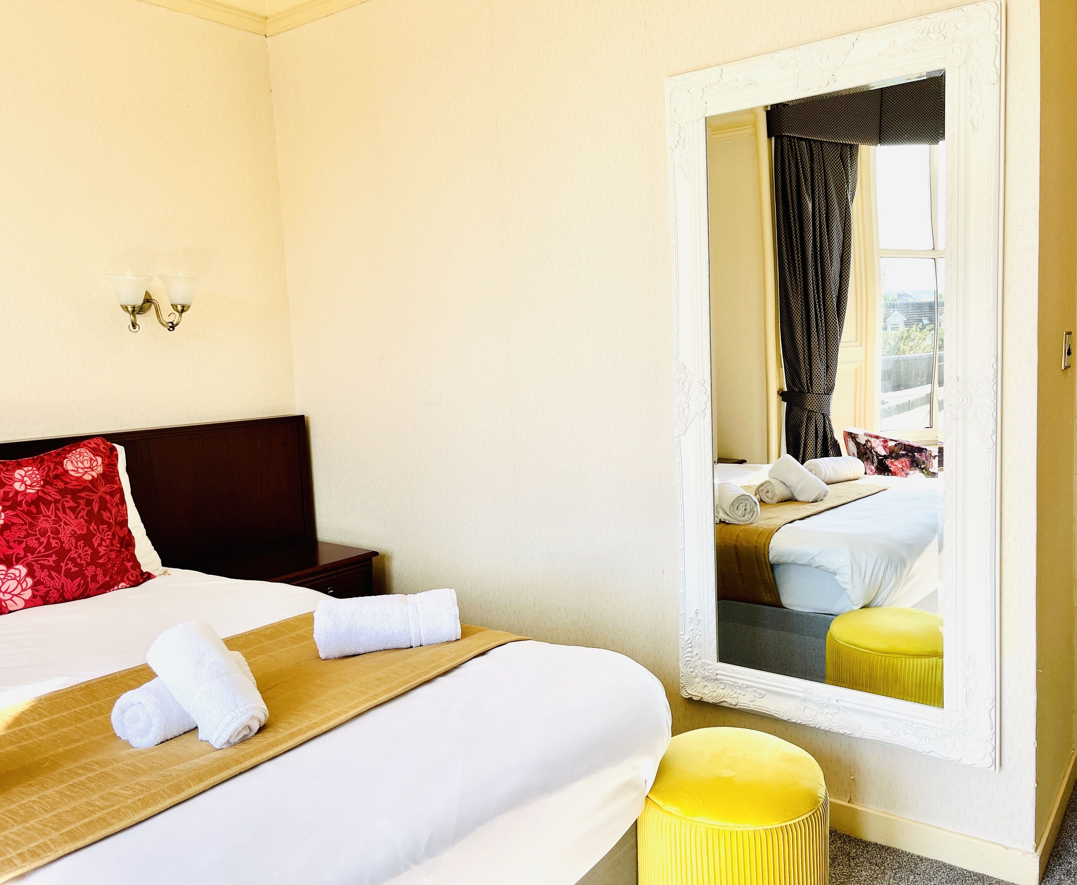 A hotel room with a large bed, a wooden headboard, a mirror, and a yellow ottoman.