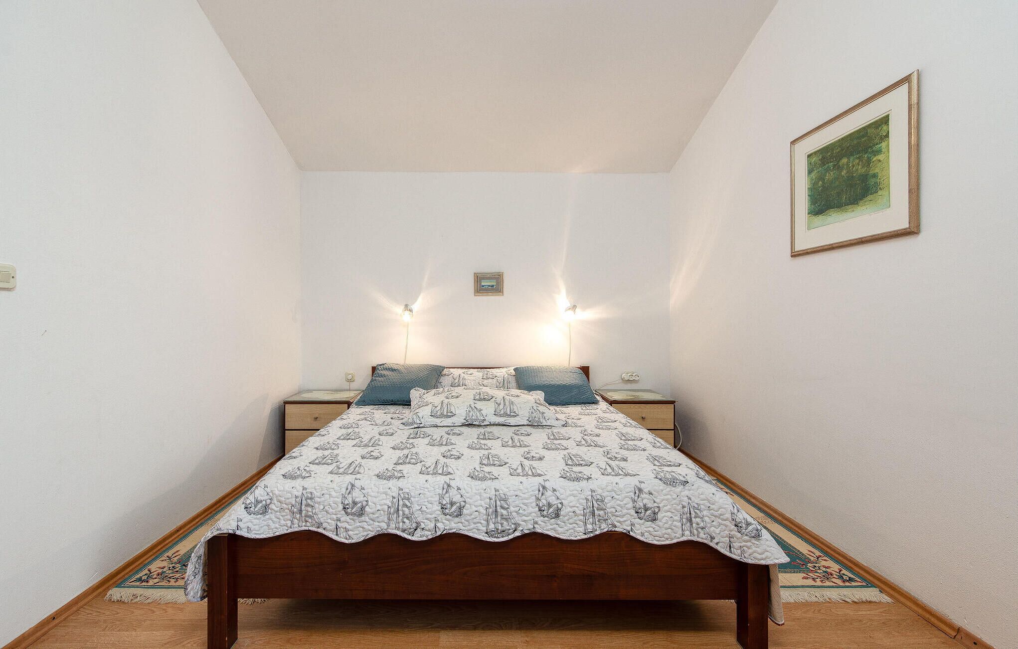 A bedroom with a wooden bed, two bedside tables, a nightstand, a wall-mounted light, and a framed picture.