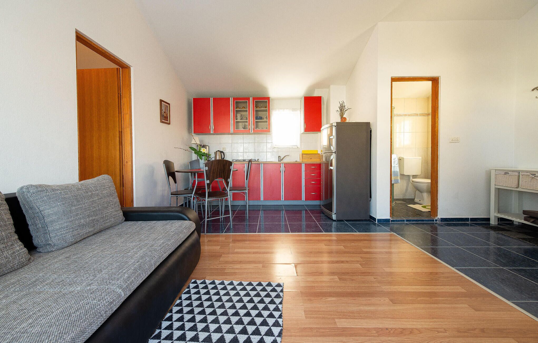 Un espace de vie compact comprenant un canapé, une table à manger avec des chaises, une cuisine avec des meubles rouges et une salle de bain visible à travers une porte ouverte.