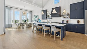 A modern kitchen with dark cabinetry, a central island with bar stools, and a dining area with a large window. - Luxe,Pet-Friendly,Lake Living@Cinnamon Shore! Game Rm, Amenities&Close Pools! (Port Aransas)