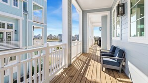 A wooden deck with white railings, a seating area with two chairs, and a view of a body of water and buildings in the distance. - Luxe,Pet-Friendly,Lake Living@Cinnamon Shore! Game Rm, Amenities&Close Pools! (Port Aransas)