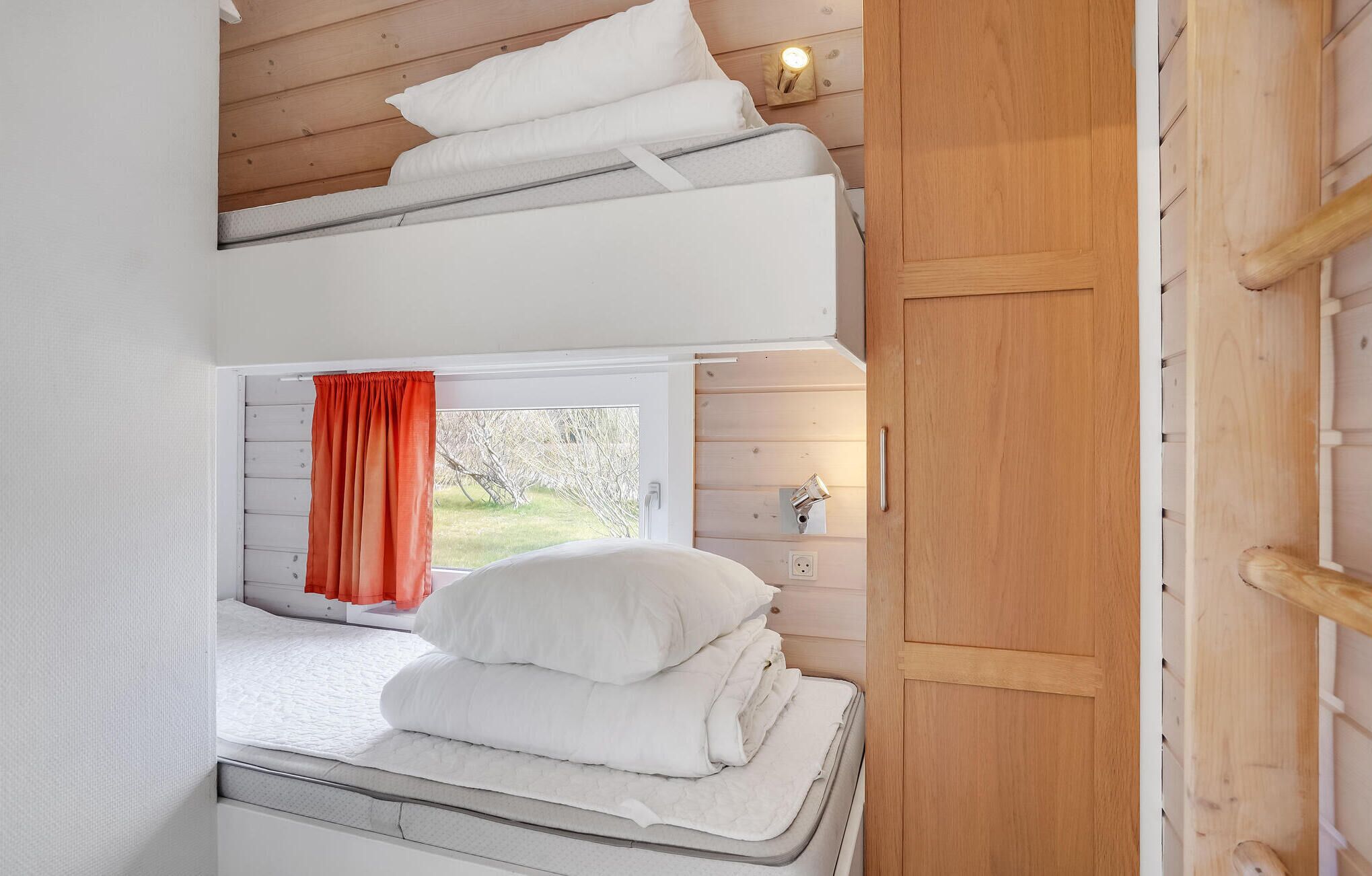 A bunk bed with white bedding, a window with a red curtain, and a wooden wall with a light switch.