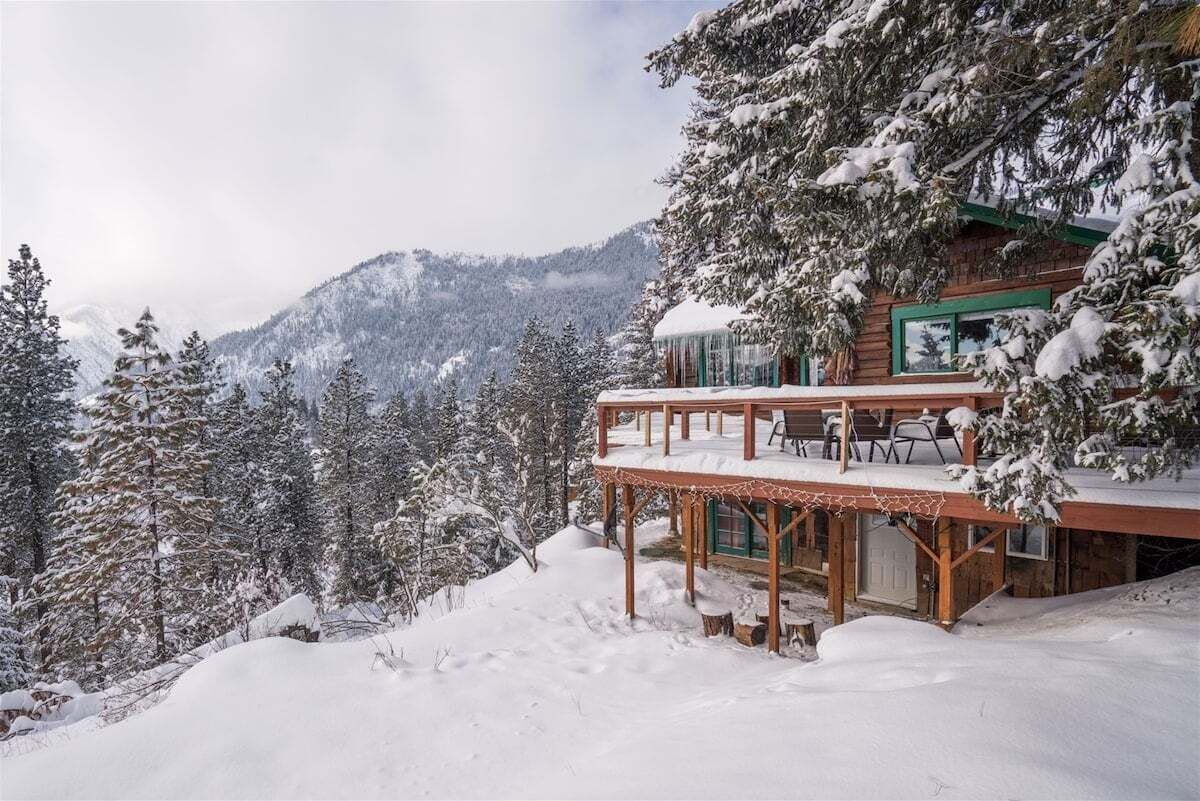 A wooden cabin with a balcony, surrounded by snow-covered trees and mountains.