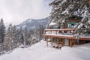 A wooden cabin with a balcony, surrounded by snow-covered trees and mountains.
