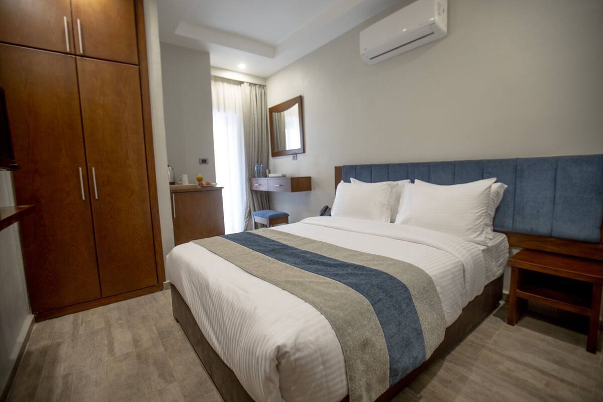 A hotel room with a bed, wooden wardrobe, bedside table, and an air conditioning unit.