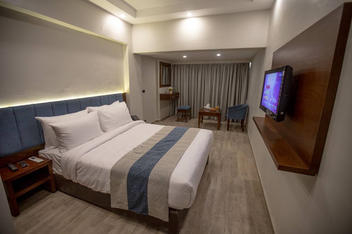 A hotel room with a large bed, bedside tables, a TV mounted on the wall, and a seating area with a chair and table.