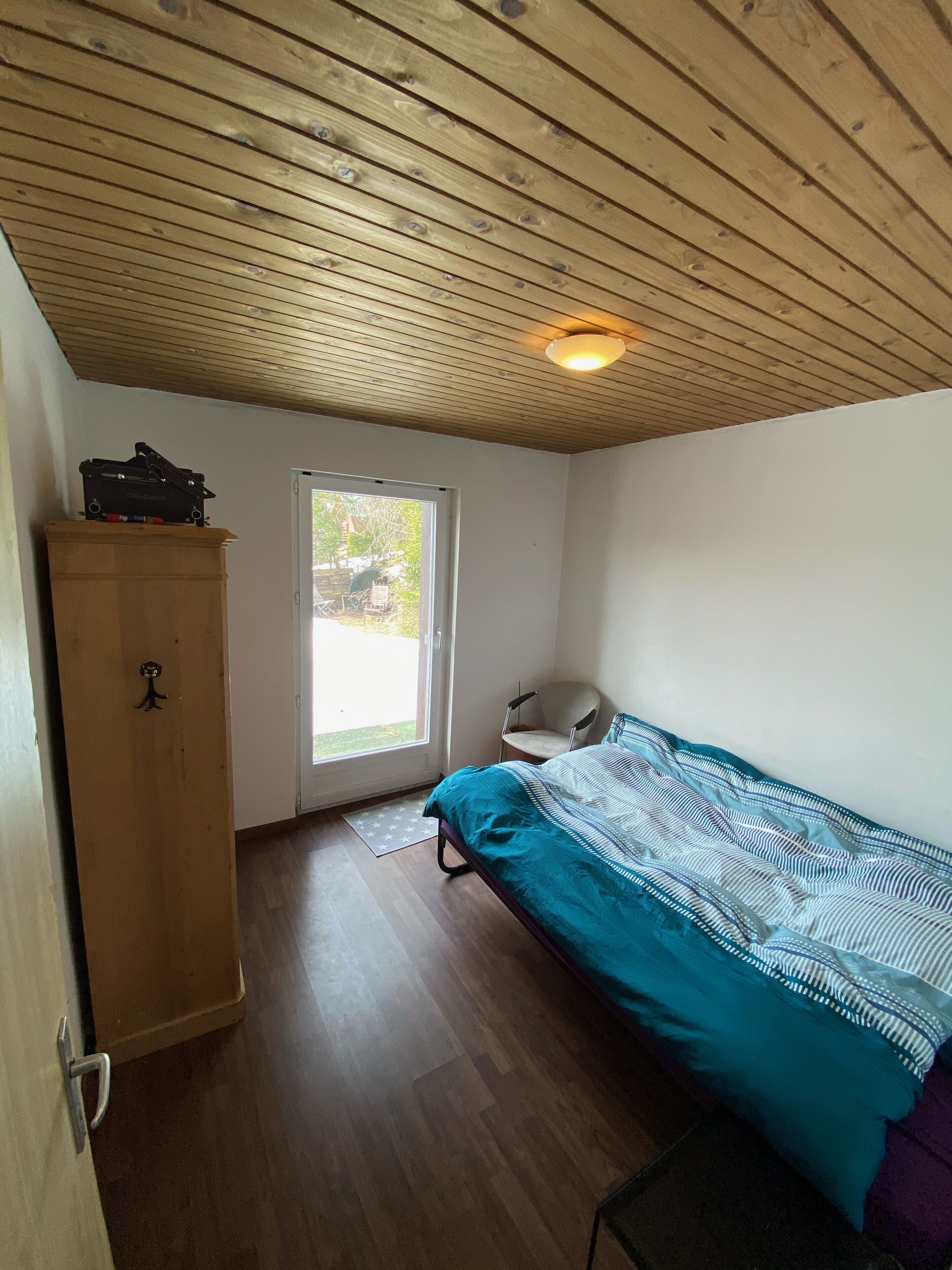 Een klein appartement met één kamer, een houten plafond, een bed met streepjesbeddengoed, een houten kledingkast en een stoel.