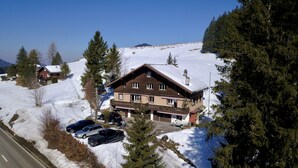 A chalet-style building with a snow-covered roof, surrounded by trees and a clear sky. - Apartment with garden at the foot of the Säntis (Urnäsch)