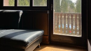 A room with a black leather sofa, wooden flooring, and a large window offering a mountain view. - Apartment with garden at the foot of the Säntis (Urnäsch)