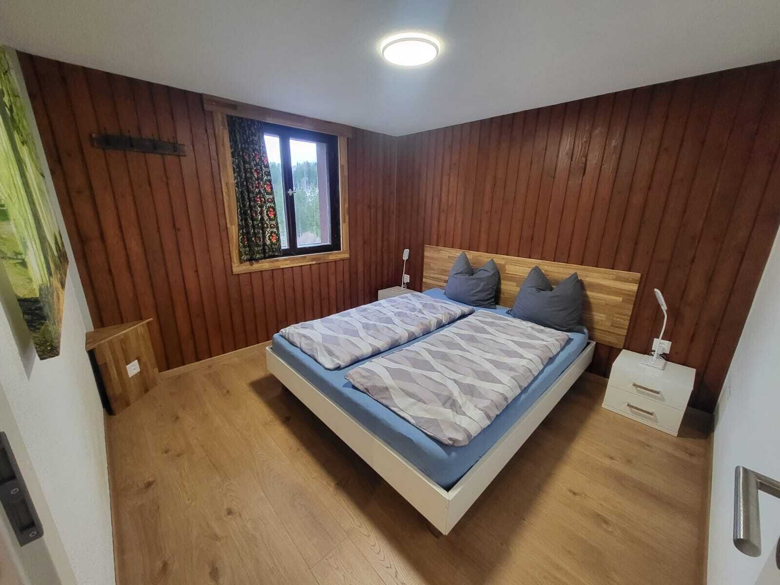 A bedroom with a wooden paneled wall, a window with curtains, a bed with blue bedding, and a bedside table with a lamp.
