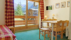A room with a wooden table and chairs, a bed with a checkered blanket, a window with a view of mountains, and a television on the wall.