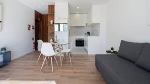 Un soggiorno moderno con un divano grigio, un'area pranzo con un tavolo in vetro e sedie bianche, e una cucina con mobili bianchi e un forno nero.
