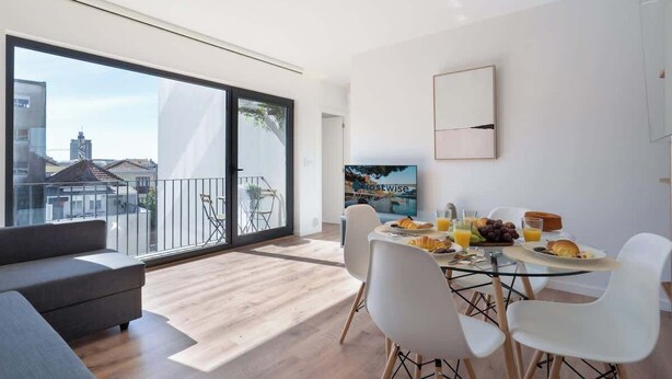 Un soggiorno moderno con una grande finestra, un'area pranzo con tavolo apparecchiato per la colazione e un balcone con vista sulla città.
