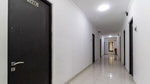 Ein langer, schmaler Gang mit weißen Wänden und einer schwarzen Tür links. Der Boden ist mit Fliesen ausgelegt, und der Gang führt zu einem Zimmer mit einem Fenster.