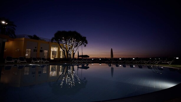 A nighttime view of a swimming pool with lounge chairs and a building with illuminated windows.