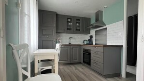 A compact kitchen with grey cabinets, a white countertop, and a dining area with a white table and chairs. - Charming house 800m from the sea and Ménéham (Kerlouan)
