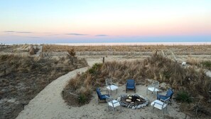 A beachside seating area with a fire pit, surrounded by chairs and a wooden fence.