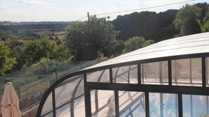 A greenhouse with a curved glass roof, surrounded by a metal fence, overlooking a rural landscape with trees and fields. - Casa Mestres - La Botiga (Pujalt)