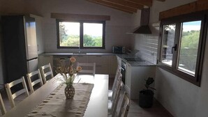 A compact kitchen with a dining area, featuring a long table, chairs, a vase with flowers, and a view through the windows. - Casa Mestres - La Botiga (Pujalt)