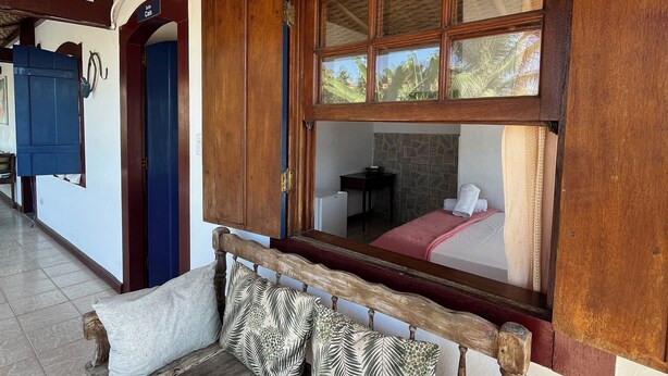 Un banco de madera con cojines estampados, un ventanal con contraventanas de madera y un suelo de baldosas.