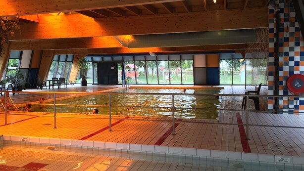 Une piscine intérieure avec un plafond en bois, un sol carrelé et une vue sur l’extérieur grâce à de grandes fenêtres.