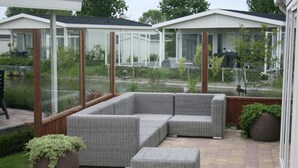 Un patio couvert, aménagé avec des meubles en osier, une plante en pot et offrant une vue sur un étang et la verdure environnante.