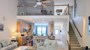 A spacious living area with a high ceiling, a kitchen with wooden cabinets, a dining area with striped chairs, and a staircase leading to an upper level. - Spacious 4BR Forest Landing Duplex - Dog Friendly, Pool & Trails! (Frankford)