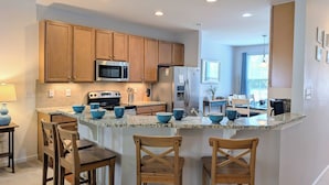 A modern kitchen with wooden cabinets, stainless steel appliances, and a granite countertop. There are three wooden chairs at the island and a dining table in the background. - Spacious 4BR Forest Landing Duplex - Dog Friendly, Pool & Trails! (Frankford)