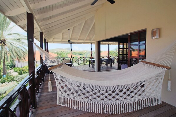 Relax in a serene outdoor space with a hammock, ceiling fans, and a wooden deck overlooking lush greenery.