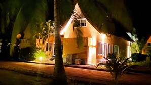Un edificio de dos pisos con techo inclinado, probablemente un hotel o resort, rodeado de palmeras e iluminado con luces exteriores.