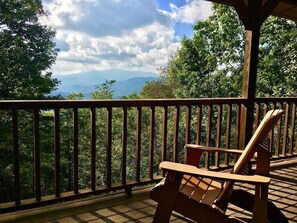 Una terraza de madera con una silla y barandilla, con vista a un área boscosa y montañas al fondo.