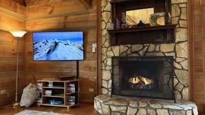 A cozy log cabin interior with a stone fireplace, a mounted television, and a bookshelf. - O'Connors Retreat - Blowing Rock, Hot tub Secluded acreage, fabulous views (Lenoir)