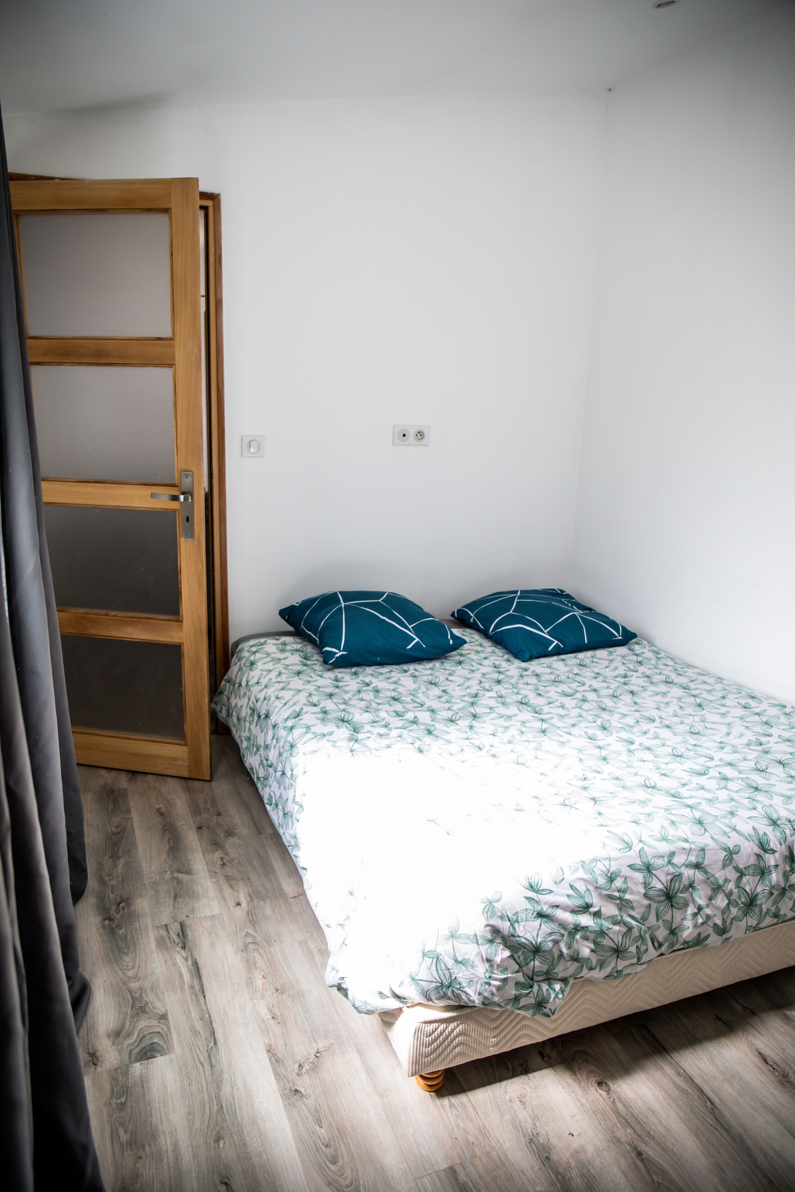 A small, single-bed room with a wooden floor, a white wall, and a wooden door.