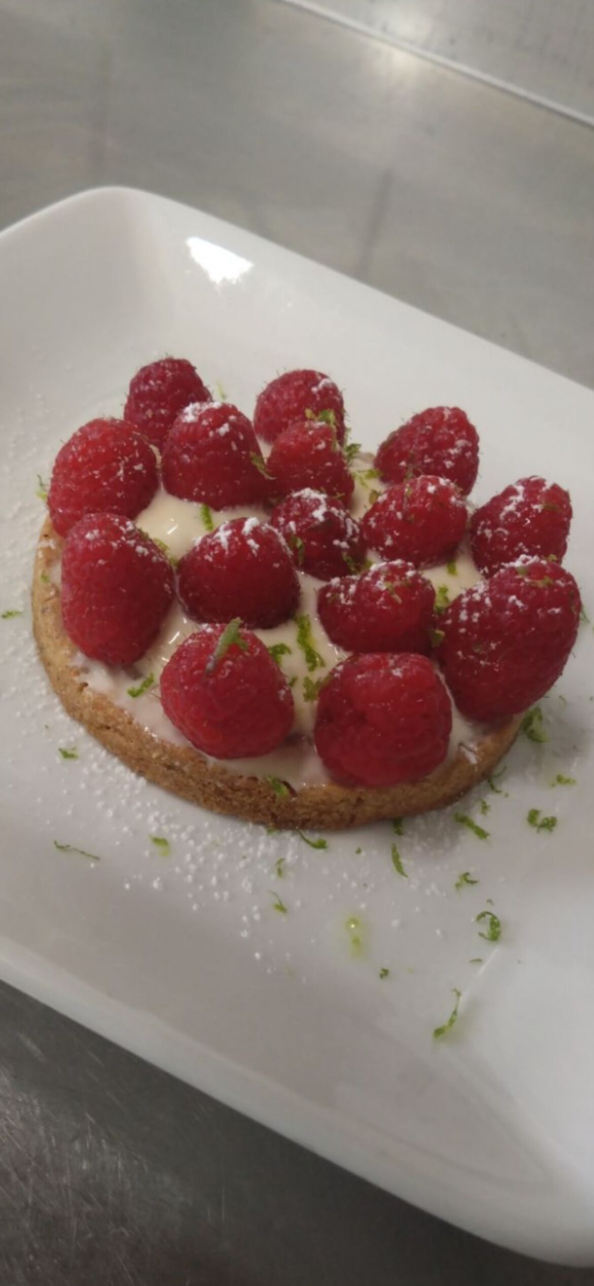 A white rectangular plate with a round pastry topped with raspberries and a white cream, garnished with green herbs.