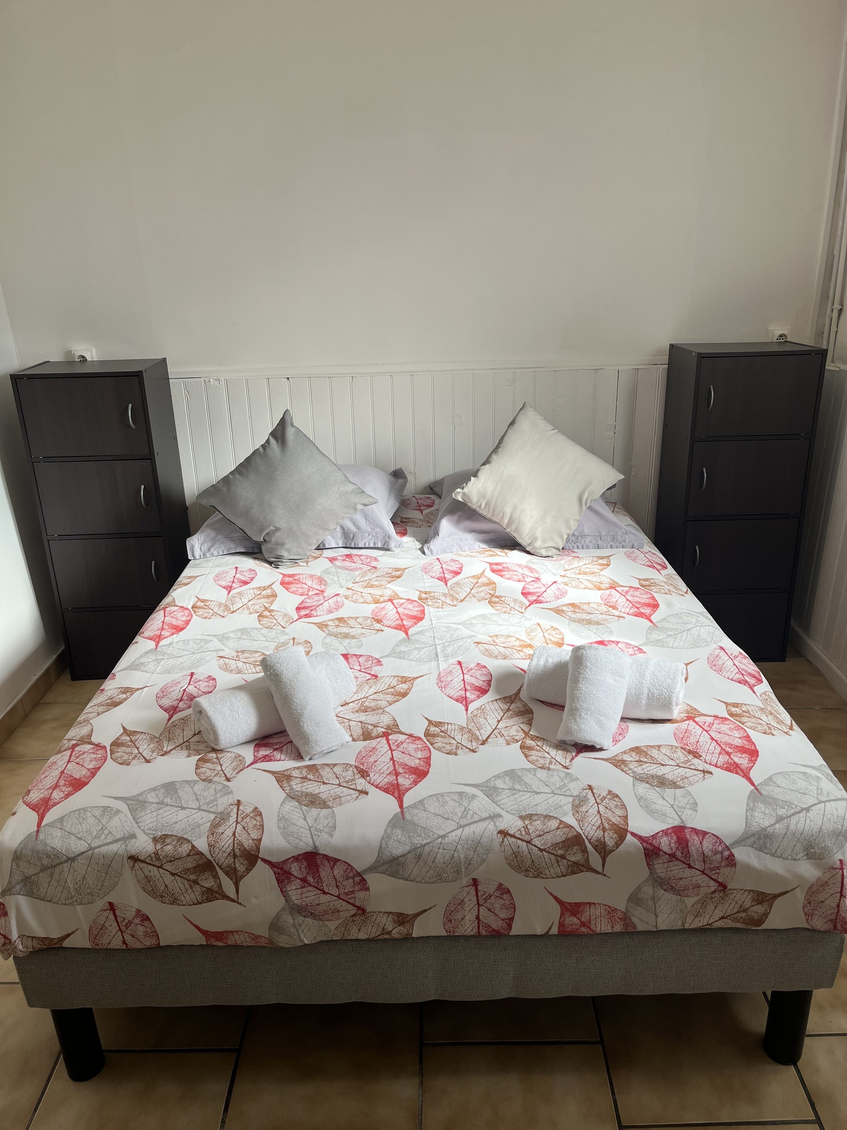 Una cama con una colcha estampada con motivos de hojas, dos almohadas y dos mesitas de noche oscuras.