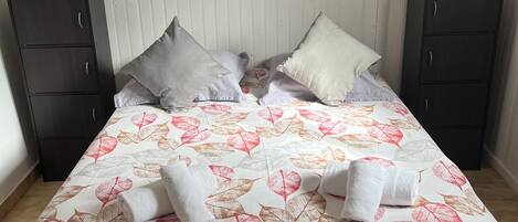 Una cama con una colcha estampada con motivos de hojas, dos almohadas y dos mesitas de noche oscuras.
