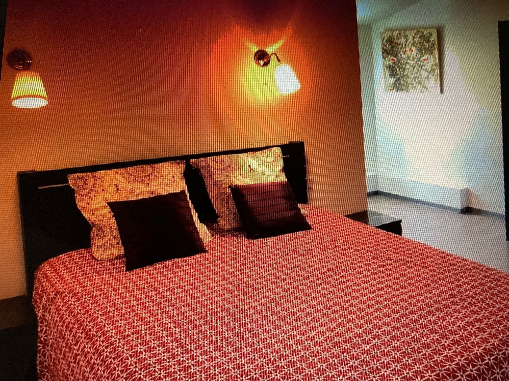 Habitación de hotel con colcha estampada, dos almohadas negras, cabecero y lámparas fijadas a la pared.