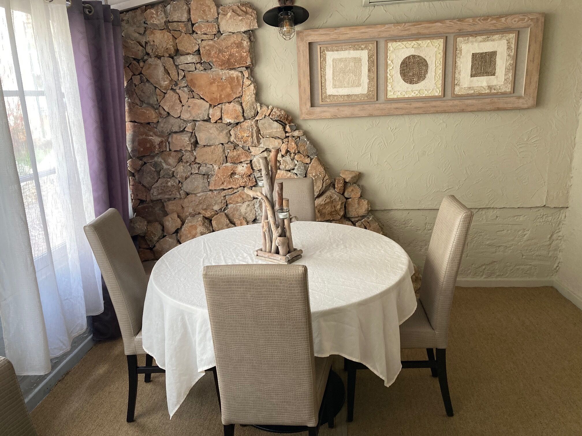 A dining area with a stone wall, a round table with a white tablecloth, and three chairs.