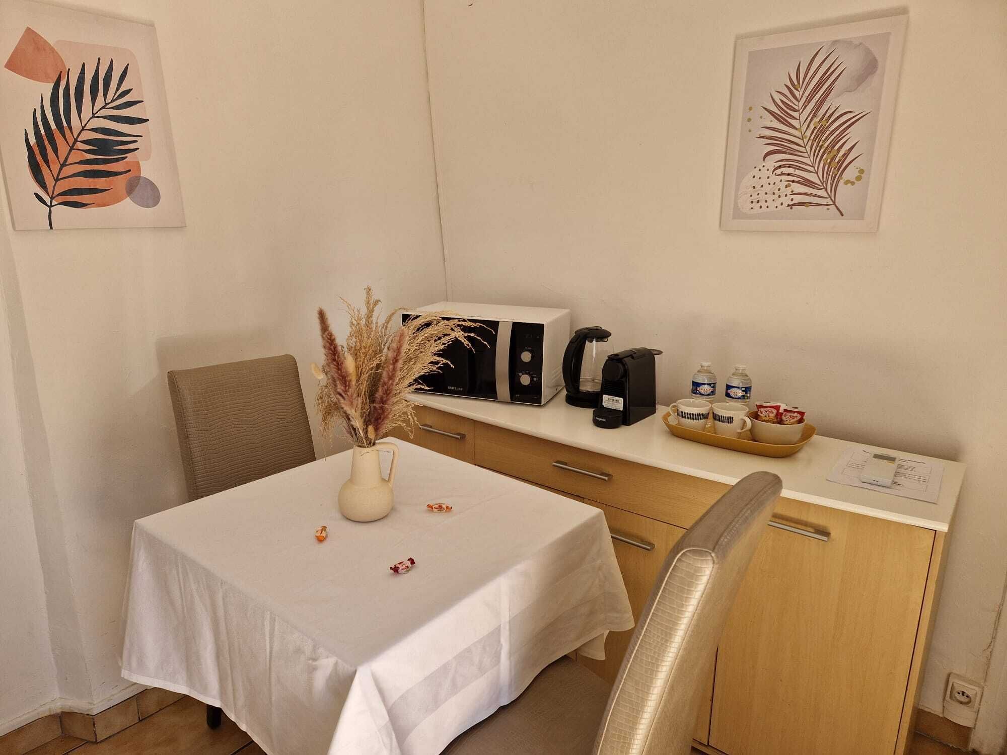 Habitación de hotel con una mesa blanca, un jarrón con plumas de plumas de pampa, una cafetera, un microondas y un mueble con una bandeja de tazas de café.
