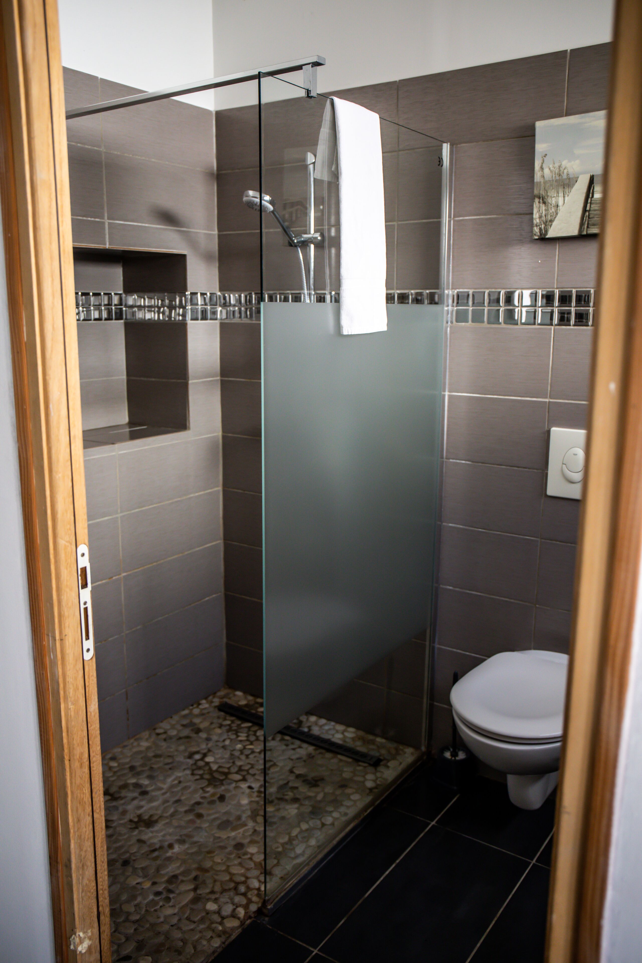 Un box doccia in vetro, un pavimento piastrellato, un WC bianco e un piccolo ripiano con articoli da toilette.