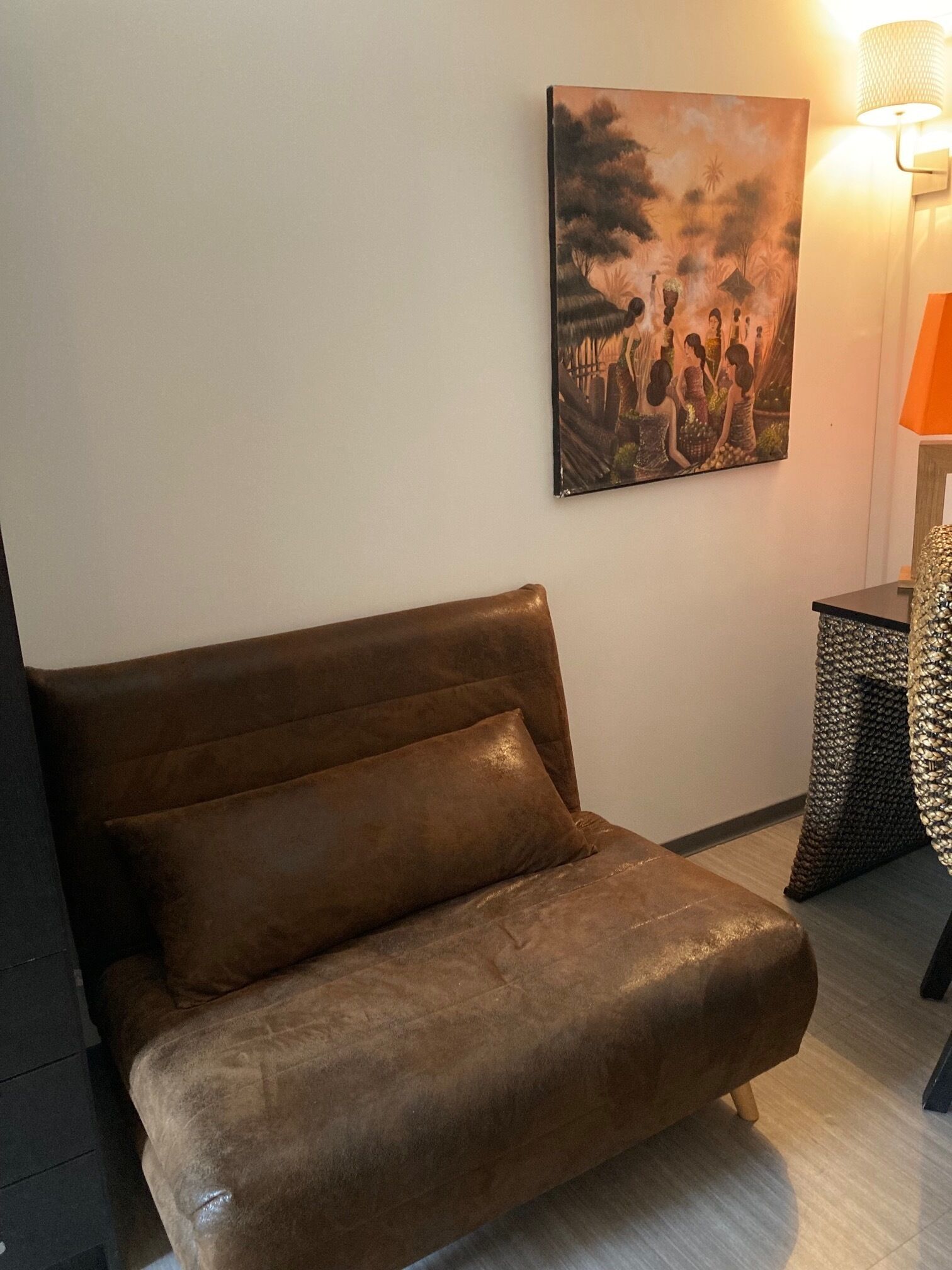 A brown leather sofa with a matching pillow in a room with a painting on the wall and a lamp on a side table.