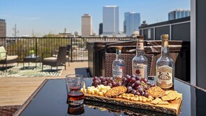 Una mesa de cristal con una selección de bocadillos, como uvas, nueces y galletas, y tres botellas de whisky bourbon en un balcón con vistas a la ciudad.